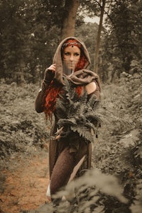 a red haired woman in a cloak standing in the woods