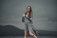 a woman in a gray dress standing in a field with a cloudy sky